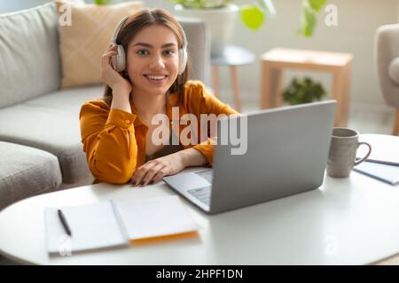 Portrait of cheery young Caucasian woman in wireless headphones using laptop pc, smiling at camera indoors Stock Photo