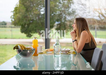 Young woman having breakfast in the living room of her house looking away Stock Photo