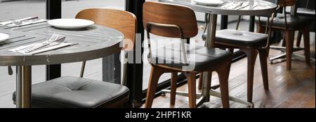 round gray plank table with stainless steel base and retro wood chair with leather cushion on old wooden floor near black frame window Stock Photo
