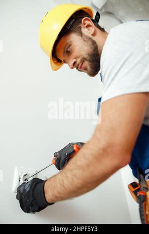 Male worker, professional electrician in hard hat and protective gloves using screwdriver while installing electrical socket outlet Stock Photo