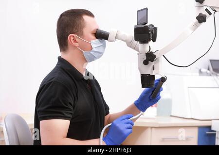 male stomatologist in protective gloves and mask on workplace, looking in dental microscope, Handsome, dentist wearing in uniform. Healthy teeth and m Stock Photo