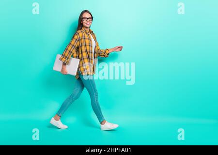 Full length profile photo of teacher young brunette lady go hold laptop wear eyesight shirt jeans sneakers isolated on teal background Stock Photo