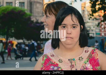 portrait of young latin caucasian woman looking at the camera and her mom behind her, standing in a plaza during sunset. lifestyle and disabled people Stock Photo