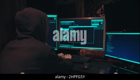 Hacker in a hood sits in front of computer screens and hacks databases Stock Photo