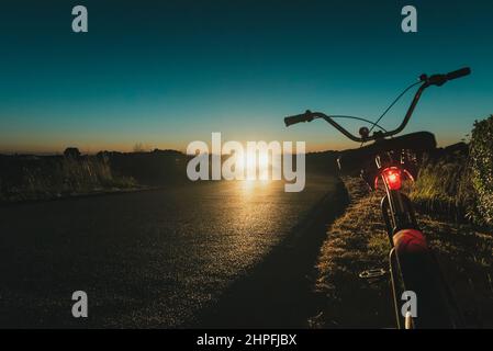 Bike on the low side of the road with red light on crossing car head light approaching by nigh Stock Photo
