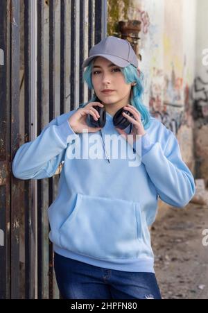 Teenager wearing light blue oversize hoodie and baseball cap looking into camera. Blue haired teen girl with wireless headphones stays outdoor near a Stock Photo