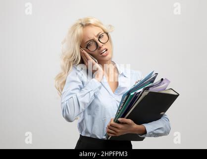 Tired young office worker with heap of folders talking on cellphone, having a lot of work, standing on light background Stock Photo