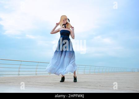 Caucasian beautiful stylish blonde sad woman in blue dress and in straw hat standing by the sea Stock Photo