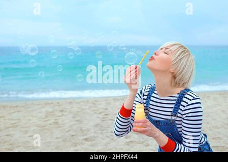 portrait of beautiful young pretty, happy, cute, blonde woman in striped shirt, denim overalls with soap bubbles by the sea Stock Photo