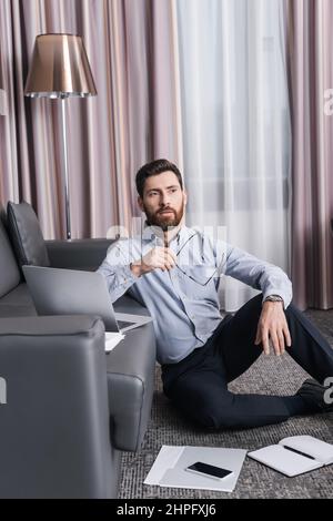pensive and bearded man holding eyeglasses and thinking near gadgets in hotel room Stock Photo
