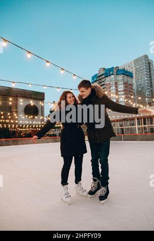 A young couple in love skating on a skating rink, arms apart, look at each other and smile. Stock Photo