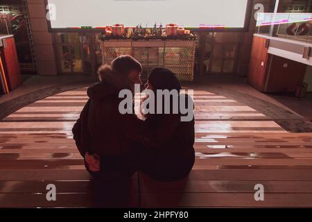 A young couple in love is sitting in a street cinema and looking at each other, hugging and kissing against the background of the screen. Stock Photo