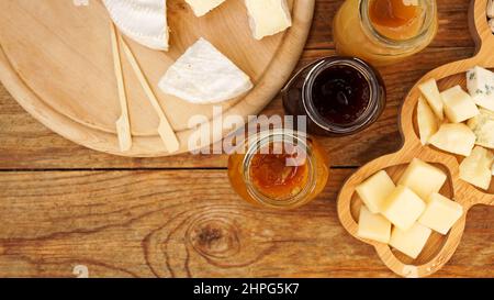 Jars of homemade jams in glass jars and a variety of cheeses on a wooden table. Cheese plate for appetizers. Top view Stock Photo