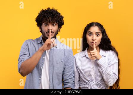 Dont say a word. Indian man and woman keeping fingers on lips, making silence sign and looking at camera, young couple standing isolated on yellow bac Stock Photo