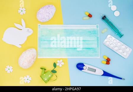 Colorful eggs, decorations, flowers and an Easter Bunny on a yellow background. Medical mask, pills, syringe, thermometer on a blue background. Holida Stock Photo