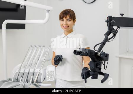 Smiling woman dentist standing in stomatology clinic near innovative microscope, among modern dental equipment. Portrait of female doctor - professional stomatologist, orthodontist. Stock Photo