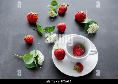 White spirea flowers and bright red strawberries in a white cup and scattered around on a black background. Summer mood background. Stock Photo