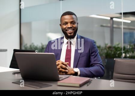 Cheerful handsome young black guy in suit businessman posing at workdesk in modern office, african american entrepreneur sitting in fornt of laptop, s Stock Photo