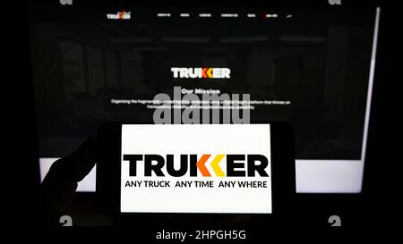 Person holding mobile phone with logo of logistics company TruKKer Holding Limited on screen in front of business web page. Focus on phone display. Stock Photo