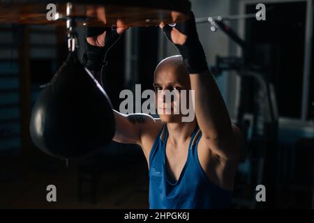 Portrait of brutal bald boxer male in boxing defence tape resting near punching bag in sport club with dark interior, looking at camera. Stock Photo