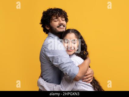 Portrait of affectionate indian couple hugging with closed eyes while standing isolated over yellow studio background Stock Photo