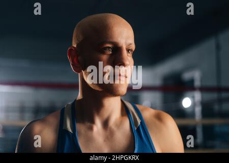 Close-up face of professional bald brutal boxer wearing sportswear standing posing in sport club with dark interior, looking away. Stock Photo