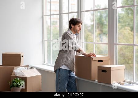 Sad tired young caucasian man in casual with cardboard boxes looks out the window Stock Photo
