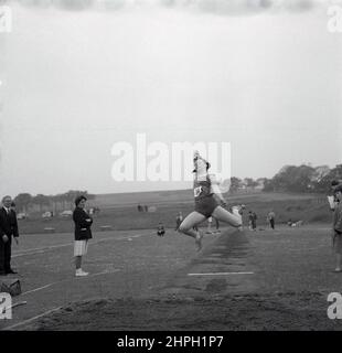 1960s, historical, a female athlete in mid-air doing the long jump, Fife, Scotland, UK. Speed, strength and agility are all needed in this track and field event, where the goal is to leap as far as possible from a takeoff point, where we see here a lady official standing opposite. A men's event in the first modern Olympics of 1896, women first took part in the Olympic long jump in 1948 in London. Stock Photo