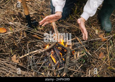 Close-up cropped shot of unrecognizable frozen tourist man warming hands over fire at outdoors on overcast cold day. Stock Photo