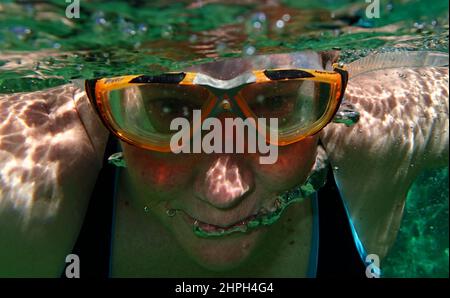 Shot from the bottom of a green tiled swimming pool, a middle aged woman (56) with wrap around goggles swims over the camera Stock Photo