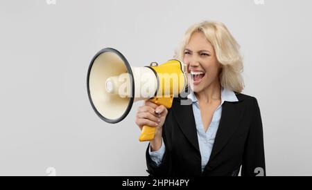 Irritated female office worker in formal wear screaming in megaphone and looking at free space, standing over light grey studio background, panorama Stock Photo