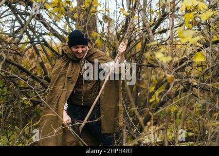 Portrait of smiling tourist male wearing raincoat tent chopping small branches with small shovel in forest on overcast cold day. Stock Photo
