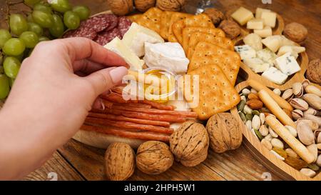 Female hand takes a breadstick and dips it in honey. Cheese and meat platter with assorted cheeses, crackers, nuts on wooden background Stock Photo