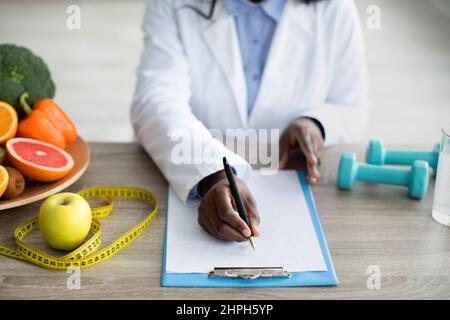 Cropped view of black female dietitian planning meal for client, writing in clipboard, sitting at desk at clinic Stock Photo