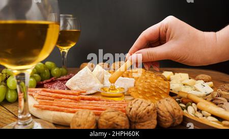 Female hand takes a breadstick and dips it in honey. Cheese and meat platter with assorted cheeses, crackers, nuts on wooden background Stock Photo