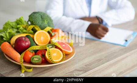 Black nutritionist writing diet plan, sitting at workplace in clinic, focus on plate with fruits and vegetables, cropped Stock Photo