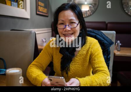 A Vietnamese woman looks at her iPhone and smiles while sitting in a cafe in Galway, Ireland. Stock Photo
