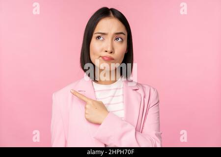 Doubtful business woman, sulking and grimacing while pointing, looking left at banner with skeptical face expression, posing against pink background Stock Photo