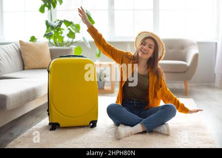 Millennial Caucasian woman making plane gesture near suitcase, sitting on floor at home, getting ready for abroad trip Stock Photo