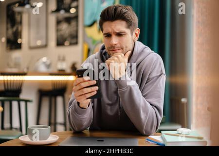 Pensive young brunette man holding mobile phone while sitting at the cafe table indoors Stock Photo