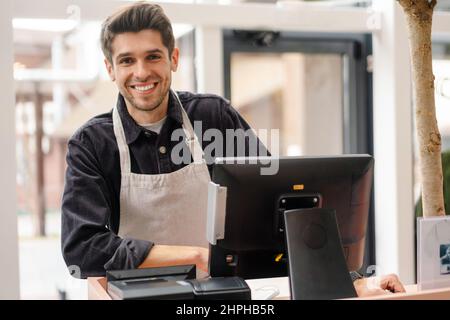 Smiling young man in apron standing at the cash register indoors Stock Photo