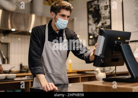 Young man in apron wearing protective medical mask using cash register in coffee shop Stock Photo