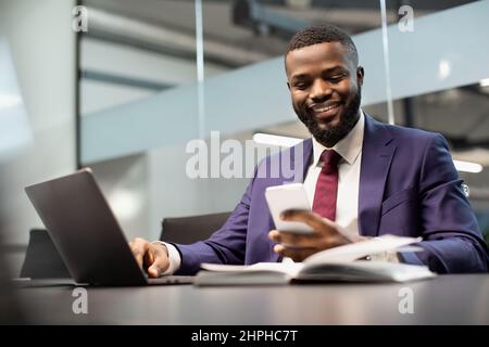 Cheerful black manager using smartphone while working on laptop Stock Photo