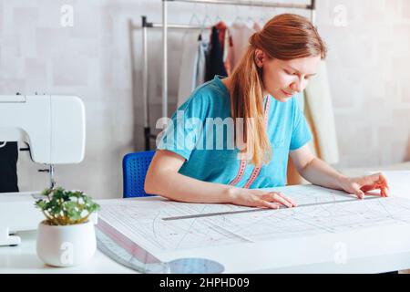 A female tailor makes a pattern . Young smiling woman fashion designer working over desk on a sewing machine. Dressmaker making a garment in her workp Stock Photo