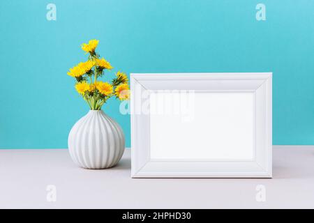 White picture frame mockup with wild yellow dandelion flowers in vase. Empty frame mock up for presentation design. White template on cian background Stock Photo