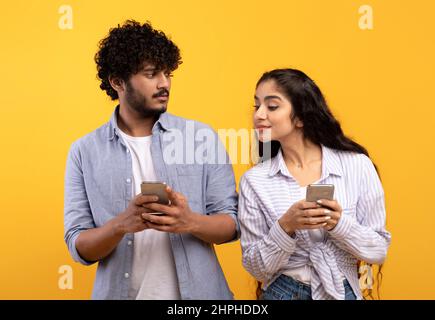 Shocked indian woman spying on her boyfriend using smartphone, texting sms or scrolling social media news feed, standing over yellow studio background Stock Photo