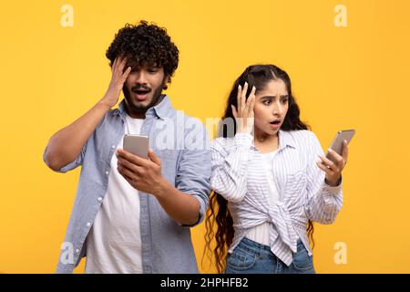 Indian woman and her boyfriend looking into their smartphones in shock and touching heads on yellow background Stock Photo