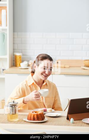 Vertical portrait of smiling woman enjoying breakfast in morning and using digital tablet in kitchen Stock Photo