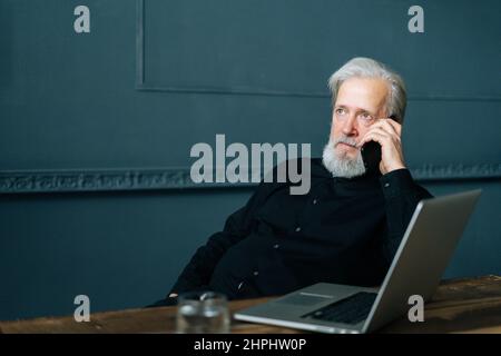 Portrait of focused gray-haired senior aged businessman talking on mobile phone sitting at wooden table with laptop computer. Stock Photo