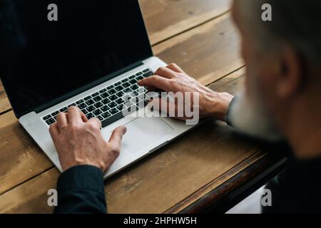 Close-up selective focus of unrecognizable bearded mature older male typing on laptop keyboard sitting at table at home office. Stock Photo